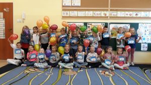 Grupa przedszkolaków w dwóch rzędach przed tablicą w sali przedszkolnej z balonami i dyplomami w dłoniach.