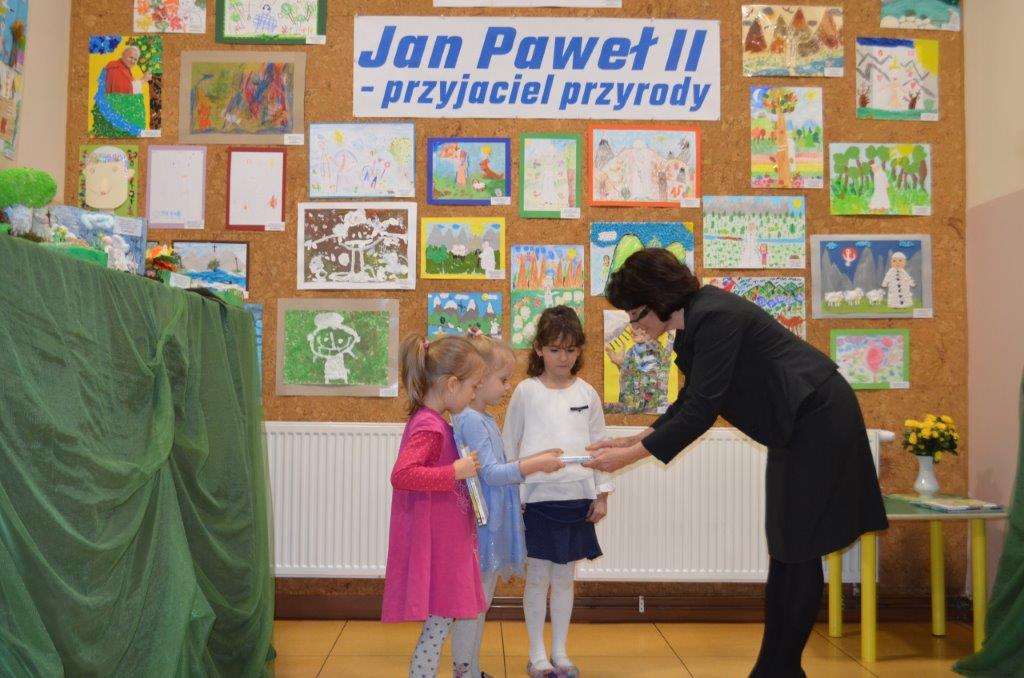 Na tle ściany z napisem „Jan Paweł II przyjaciel przyrody” stoją trzy dziewczynki, środkowa w niebieskiej sukience odbiera od Pani Dyrektor książkę.