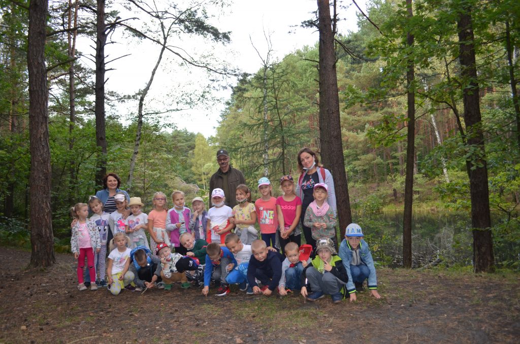 Grupa przedszkolaków z Panią Dyrektor, nauczycielem i leśniczym stoją na tle rezerwatu przyrody.
