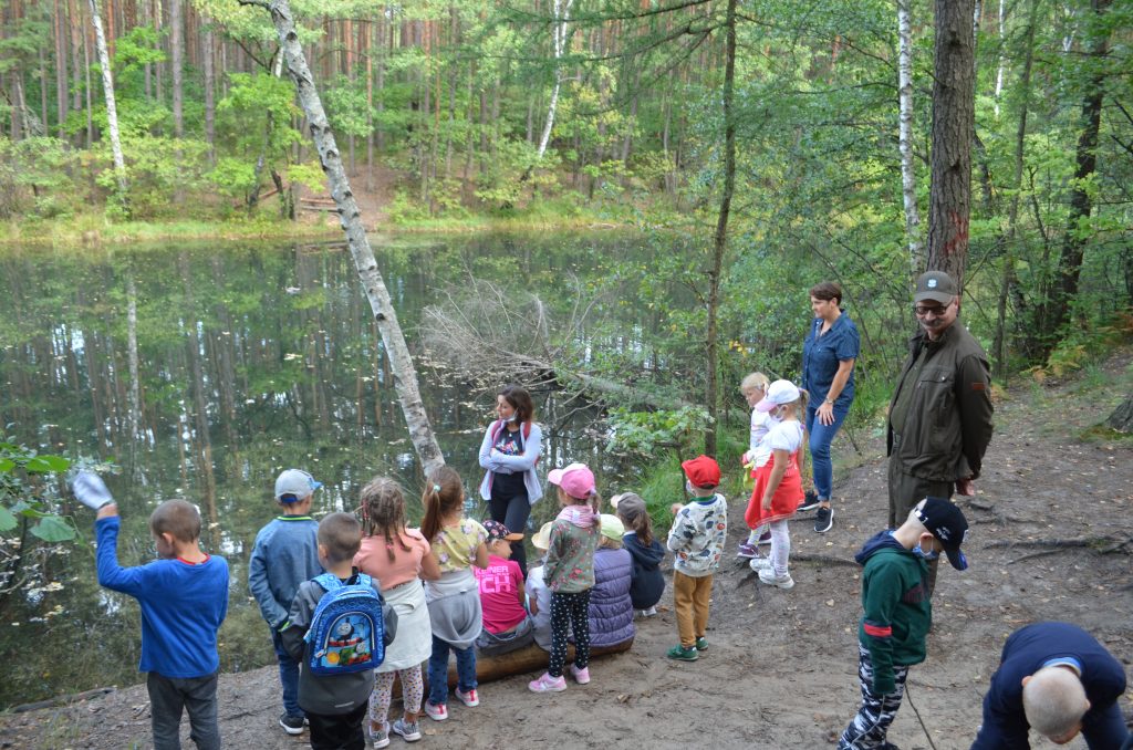 Dzieci z opiekunem stoją nad brzegiem potoku Żabnik i obserwują przyrodę.