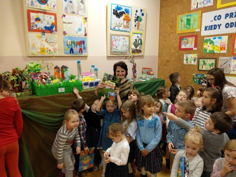 Grupa dzieci ogląda prace w Galerii. Dziewczynka w niebieskiej sukience trzyma w górze swoją książkę.