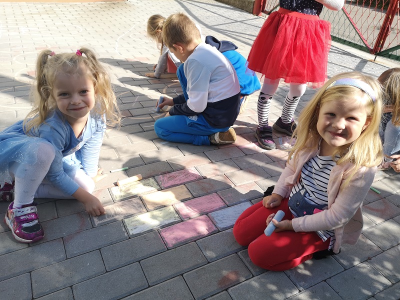 Zdjęcie przedstawia dziewczynki rysujące kolorową kredą po chodniku.