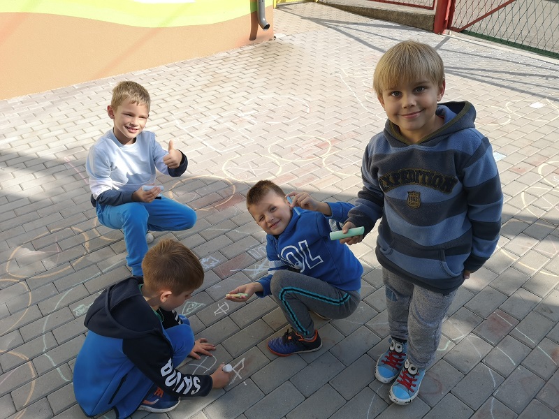 Zdjęcie przedstawia chłopców rysujące kolorową kredą po chodniku.