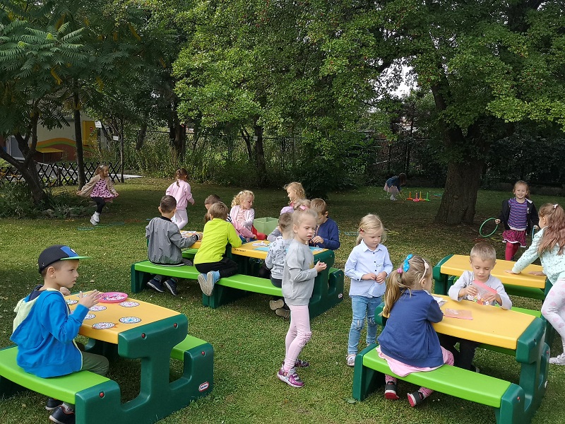 Zdjęcie przedstawia dzieci siedzące przy ogrodowych stolikach. Przedszkolaki grają w gry planszowe. Kilka dzieci uczestniczy w zabawach ruchowych na trawie.