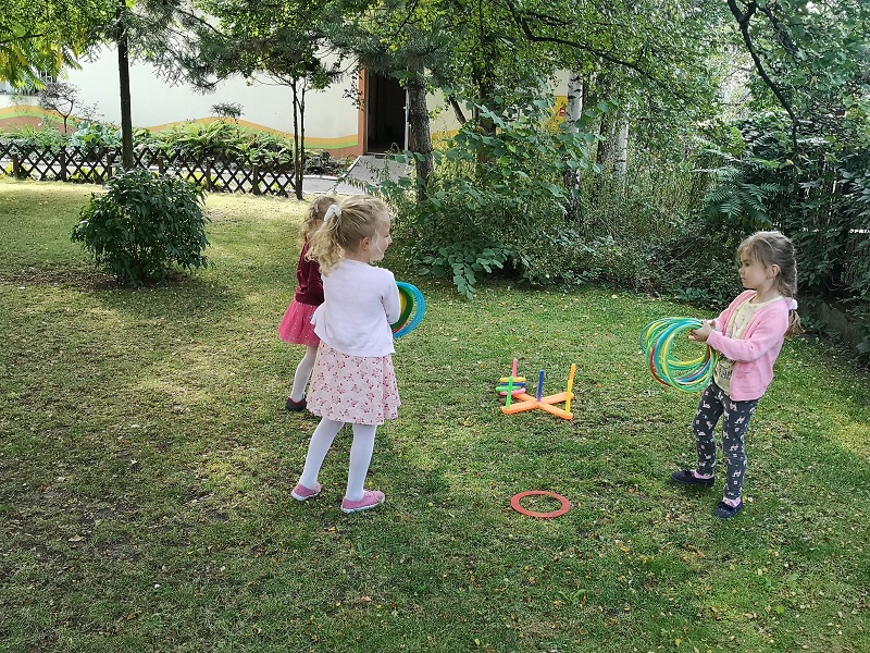 Zdjęcie przedstawia dziewczynki bawiące się w zabawę ruchową z kolorowymi obręczami – rzucanie na pachołek.