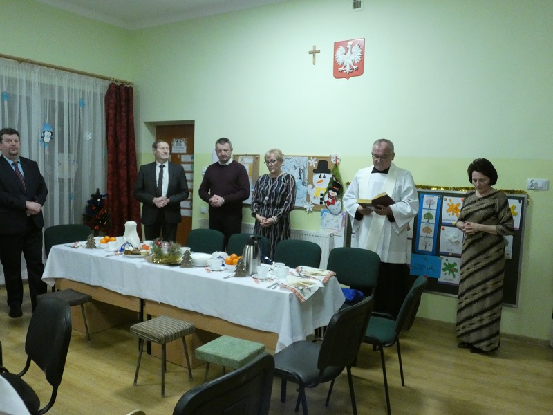 Ksiądz z otwartym modlitewnikiem i goście stoją za stołem z białym obrusem.