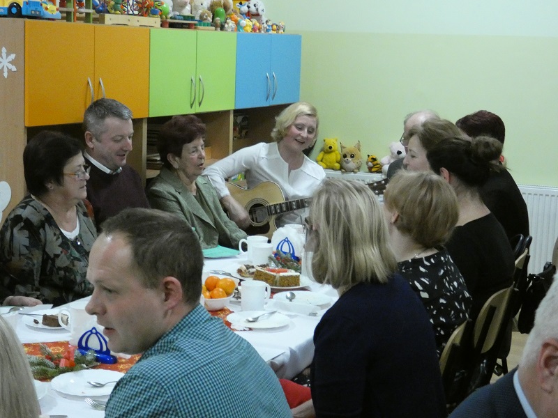Goście siedzą przy stole i śpiewają. Na wprost widać kobietę z gitara.