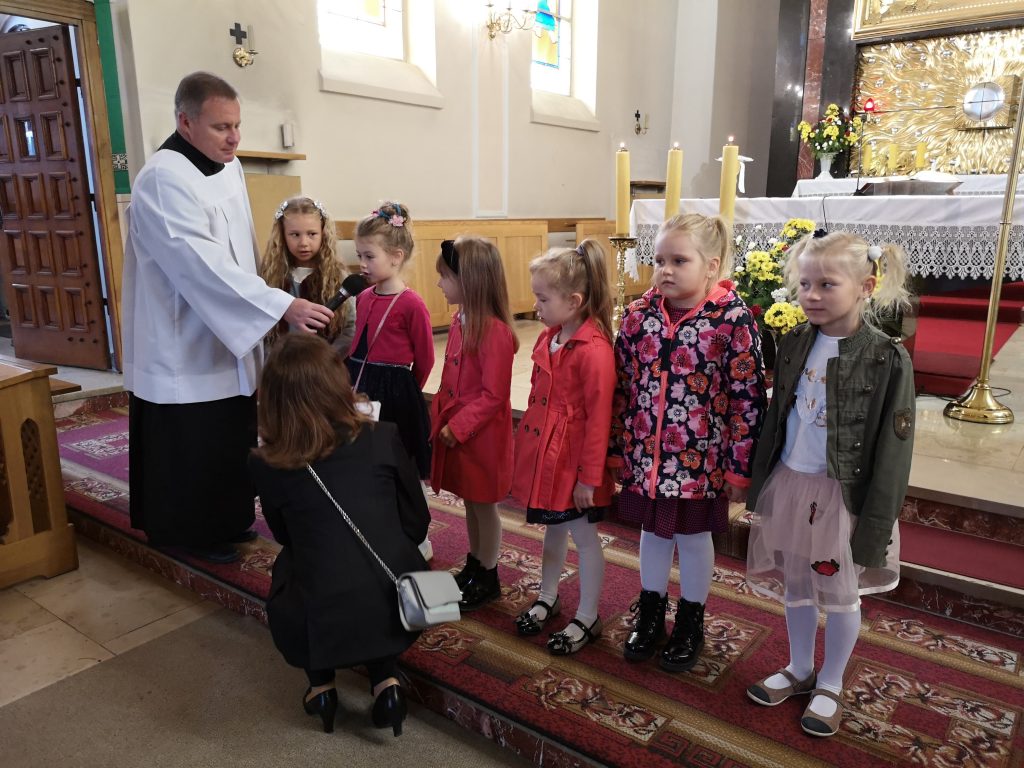 Dzieci stoją w rzędzie do modlitwy wiernych. Po lewej stronie stoi ksiądz z mikrofonem. Przed dziećmi kuca nauczyciel.