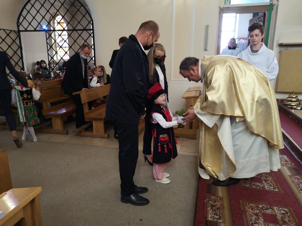 Chłopiec w stroju krakowskim składa na ręce księdza dar. Za nim stoją rodzice.