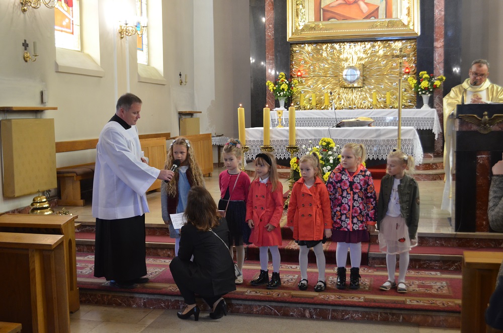 Dzieci stoją w rzędzie do modlitwy wiernych. Po lewej stronie stoi ksiądz z mikrofonem. Przed dziećmi kuca nauczyciel.