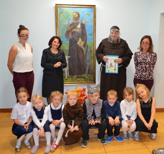 W holu przedszkola przed obrazem świętego Franciszka stoją: Pani Dyrektor, zaproszony gość ojciec Franciszkanin, nauczycielki, a przed nimi na ławce siedzą dzieci.