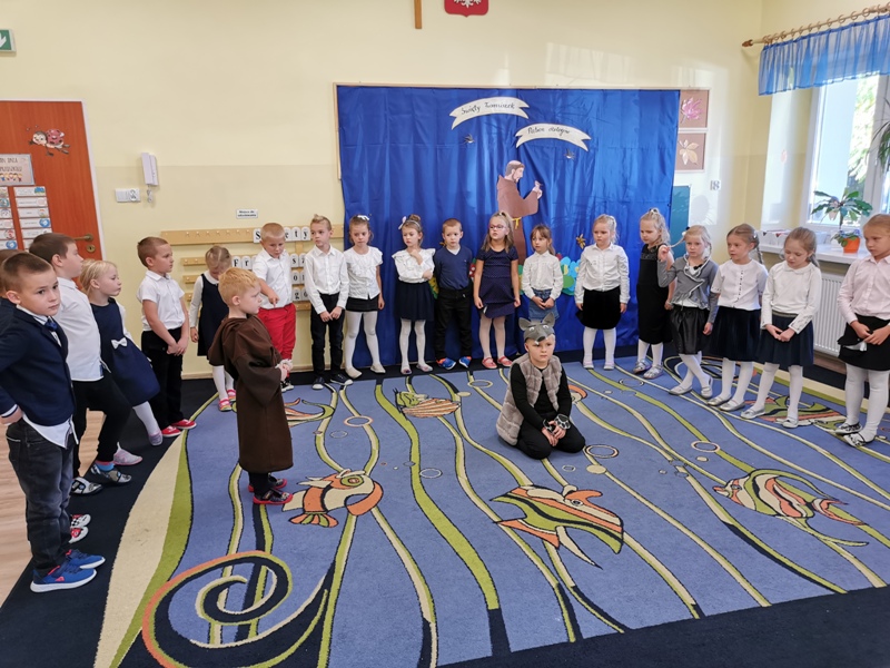 W sali przedszkolnej w półkolu stoi grupa przedszkolaków w strojach galowych oraz dwoje dzieci przebranych za świętego Franciszka i wilka.