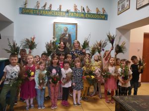 Zdjęcie grupowe pod obrazem św. Franciszka w przedszkolu z palmami wielkanocnymi.