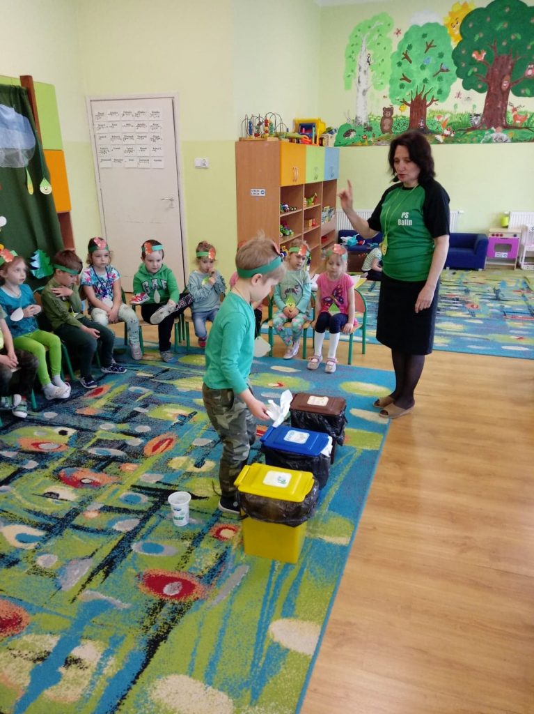 Chłopiec w zielonek koszulce wrzuca papier do niebieskiego kosza. Na drugim planie dzieci na krzesełkach i stojąca Pani Dyrektor