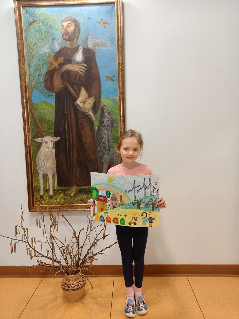 Dziewczynka stoi przed obrazem świętego Franciszka, trzymając w dłoniach pracę konkursową.
