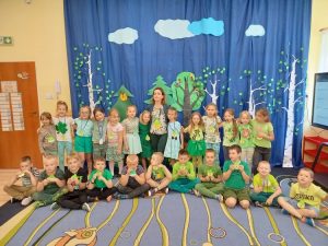 Dzieci wraz z nauczycielką w zielonych strojach, z zawieszonymi na szyi emblematami „Skrzata Ekologa”.