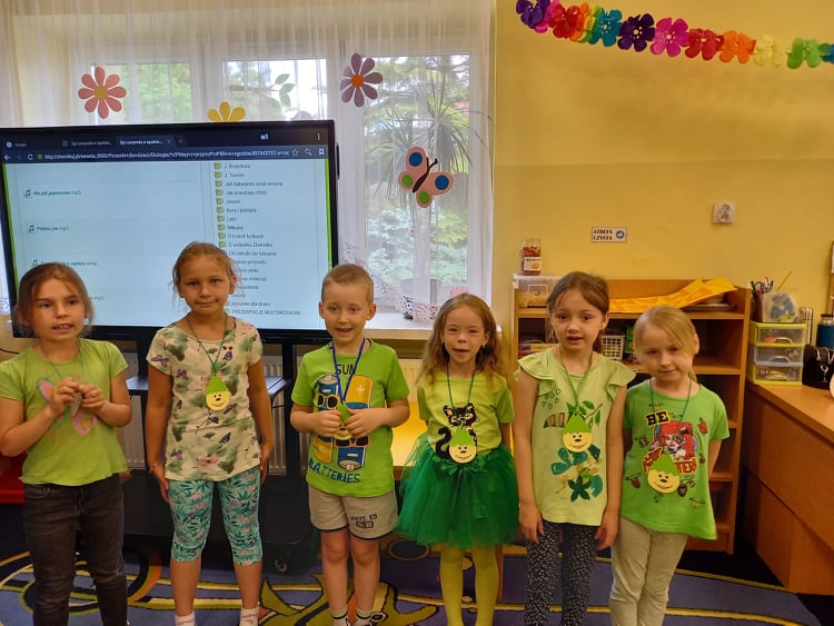 Dzieci stoją w sali przedszkolnej w zielonych strojach z emblematami Skrzata Ekologa.
