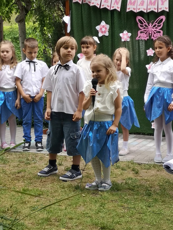 Dziewczynka stoi na środku sceny i recytuje wierszyk do mikrofonu podczas uroczystości Dnia Mamy i Taty.