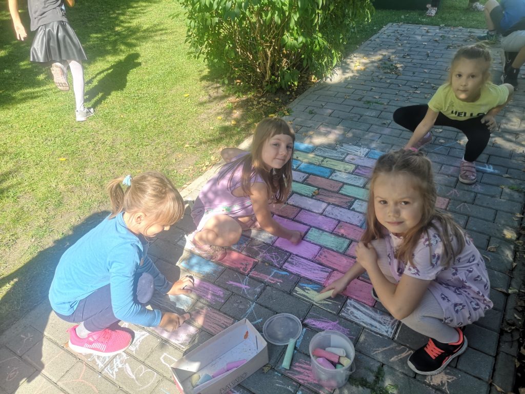 Dziewczynki malują kredą po płytkach chodnikowych.