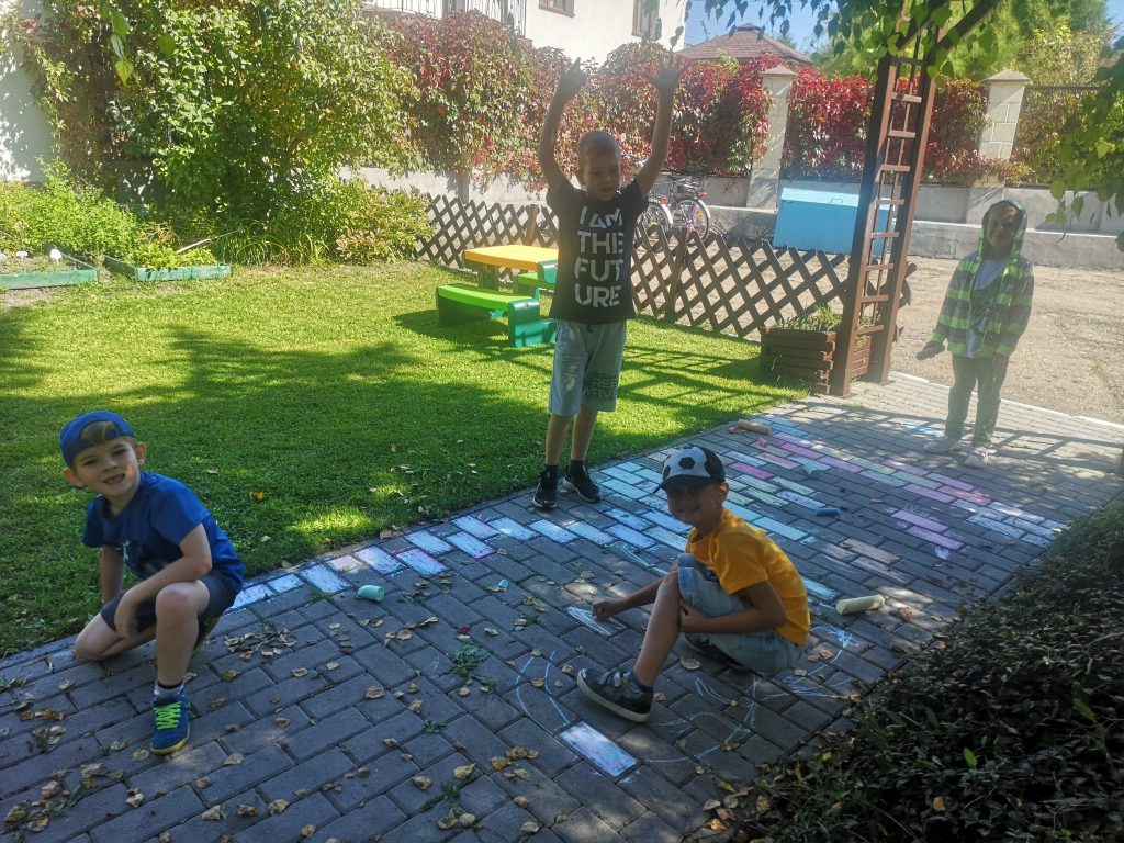Chłopcy malują kredą po płytkach chodnikowych.