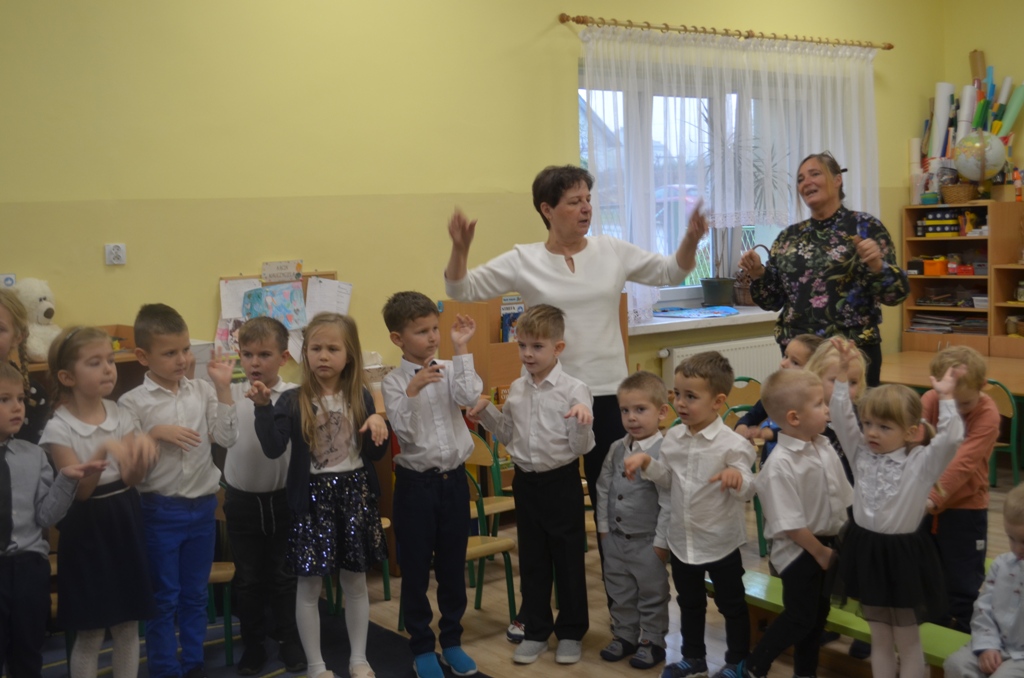 Zdjęcie przedstawia nauczyciela, pracownika oraz dzieci śpiewające hymn przedszkola.