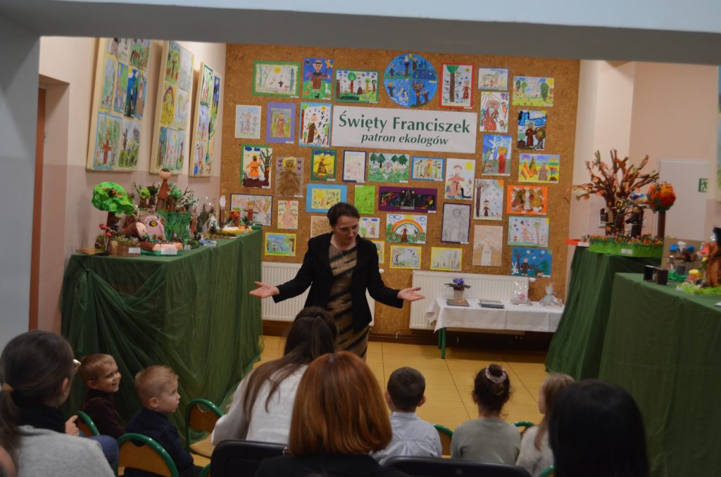 Pani Dyrektor na tle galerii dziecięcej przemawia do dzieci oraz rodziców zgromadzonych na wernisażu.
