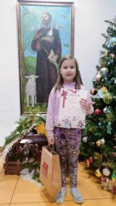 Laureatka wojewódzkiego konkursu plastycznego wraz z dyplomem i nagrodą stoi w holu przedszkola.