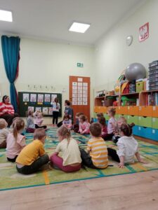 Dzieci siedzące w kole wraz z nauczycielem. Jedno dziecko przed tablicą układa odpowiednią ilość jajek w koszyczku.